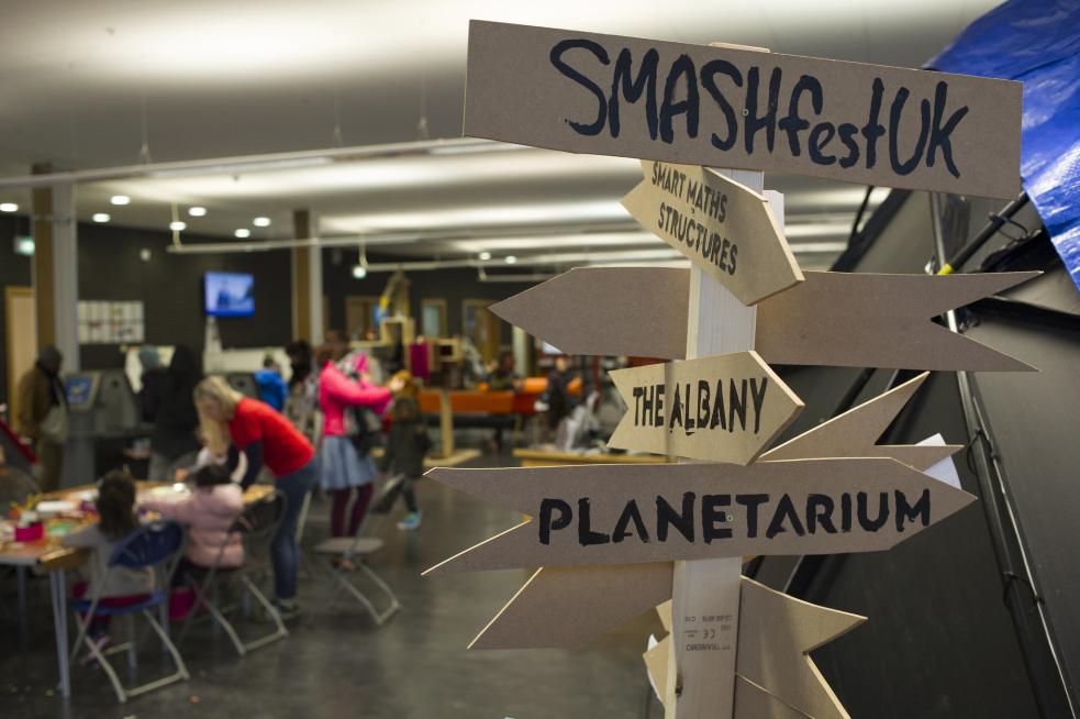 In the foreground, wooden direction signs pointing to 'SmashFestUK' and the Planetarium. In the background, children are taking part in activities at a table