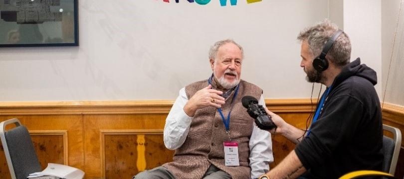 One of the Community Cast team wearing headphones and holding a microphone interviewing Budd Hall at Engage 2024, under bunting spelling out 'convening knowledge'