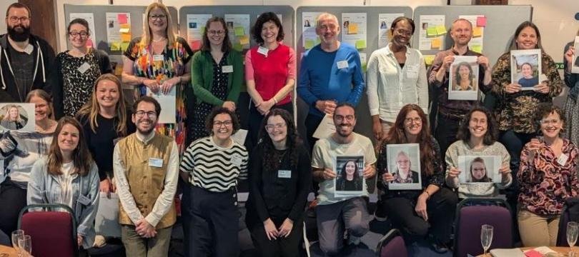 The Academy 2023_24 cohort standing/kneeling in two rows, some holding images of those not in the room