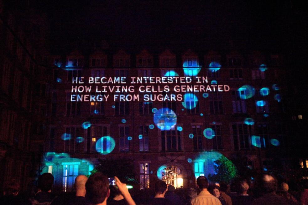 Image projected on the side of building, caption: He became interested in how living cells generated energy from sugars