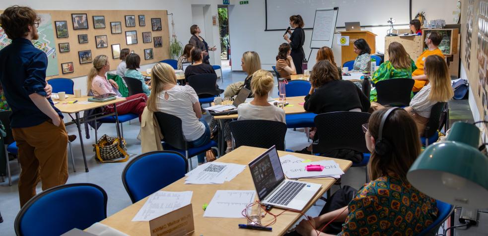 Academy 2023 - A group of around 15 people sitting around tables in a meeting room, looking to the front and listening to two people presenting in front of a screen and flip chart 