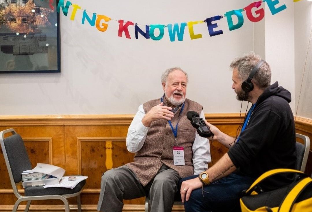 One of the Community Cast team wearing headphones and holding a microphone interviewing Budd Hall at Engage 2024, under bunting spelling out 'convening knowledge'