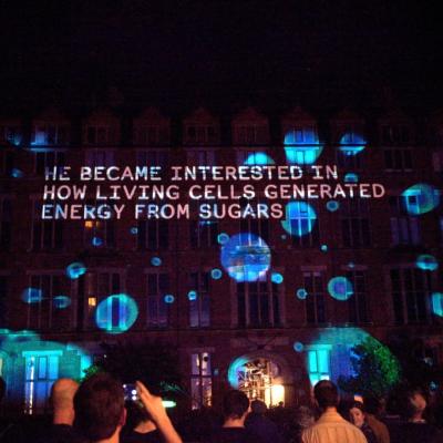Image projected on the side of building, caption: He became interested in how living cells generated energy from sugars