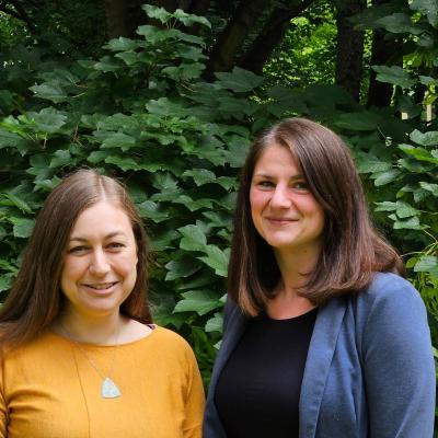 Kate and Sarah standing side by side smiling  in front of some trees 