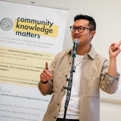 Lewis Hou presenting on stage, speaking into a microphone with an information board behind him 