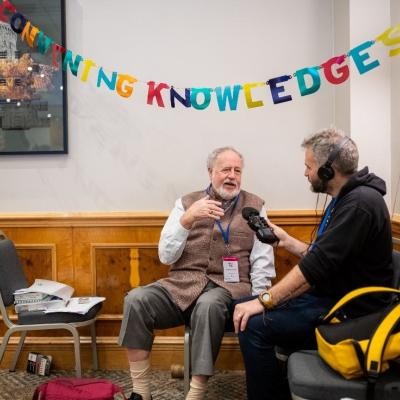 One of the Community Cast team wearing headphones and holding a microphone interviewing Budd Hall at Engage 2024, under bunting spelling out 'convening knowledge'