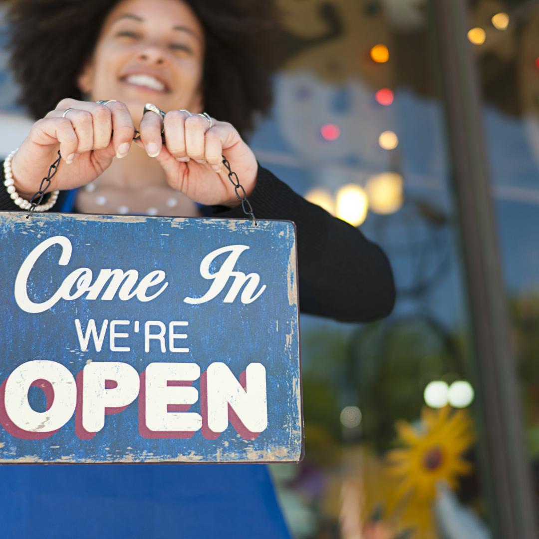 A woman smiling and holding up a shop sign that says 'come in we're open'