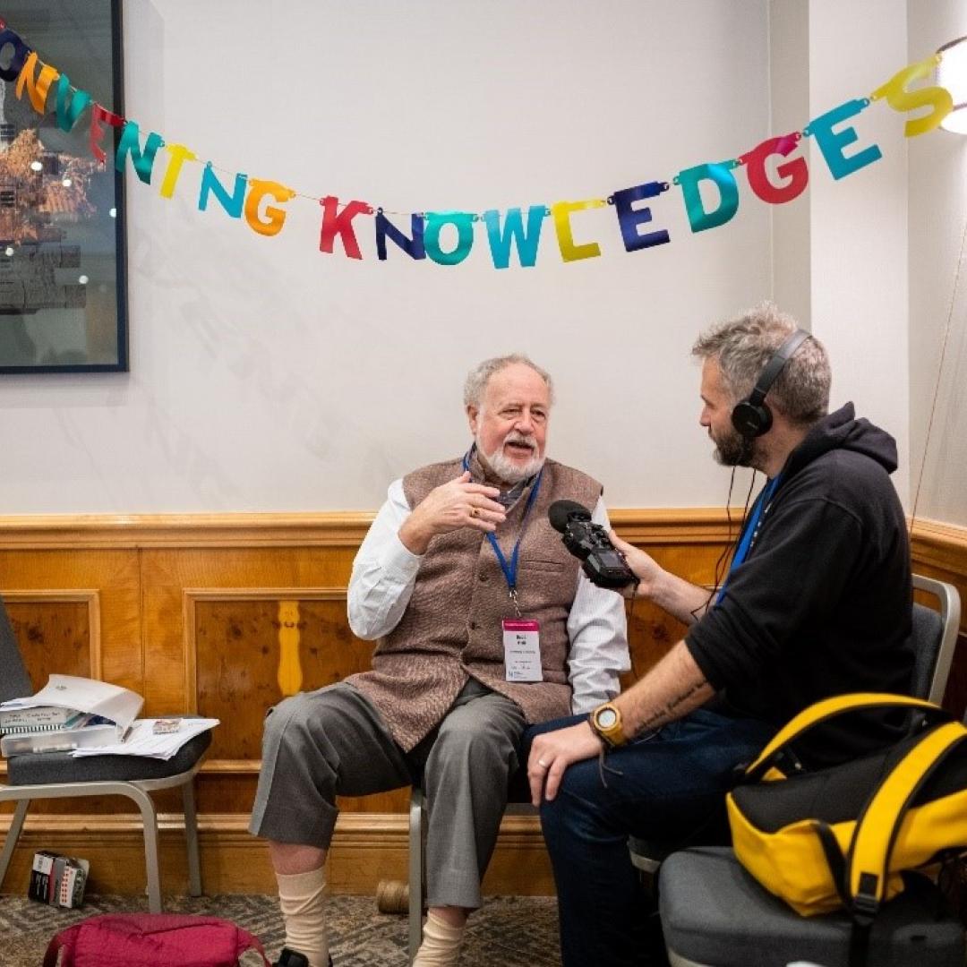 One of the Community Cast team wearing headphones and holding a microphone interviewing Budd Hall at Engage 2024, under bunting spelling out 'convening knowledge'